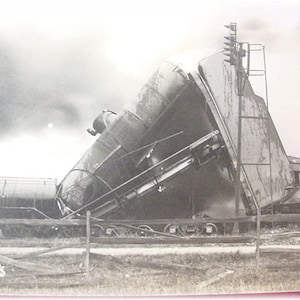 May include: A black and white photograph of a train derailment. A train car is tipped on its side, with the words "PRODU" visible on the side of the car. The train is on a track, and there is a fence in the foreground.