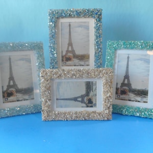 May include: Four rectangular picture frames with textured, crushed glass borders in shades of blue, teal, and silver. Each frame contains a photograph of the Eiffel Tower in Paris, displayed against a solid light blue background.