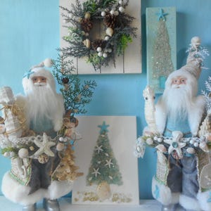 May include: Coastal Christmas decorations featuring two Santa Claus figurines with white beards and hats, adorned with seashells and starfish. A wreath and two framed Christmas trees are in the background, all against a light blue backdrop.