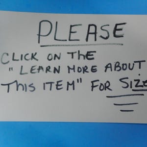 May include: A rectangular light gray sign with black handwritten text that reads: PLEASE CLICK ON THE "LEARN MORE ABOUT THIS ITEM" FOR SIZES. The text is written in capital letters with a line drawn underneath the word PLEASE and three lines below SIZES.
