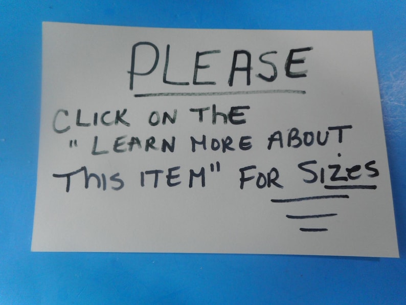 May include: A rectangular light gray sign with black handwritten text that reads: PLEASE CLICK ON THE "LEARN MORE ABOUT THIS ITEM" FOR SIZES. The text is written in capital letters with a line drawn underneath the word PLEASE and three lines below SIZES.