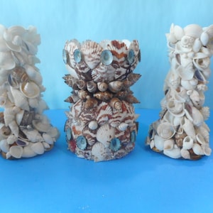 May include: Three decorative candle holders made of seashells. The center holder is brown and white with blue accents. The two outer holders are white with a variety of seashells.