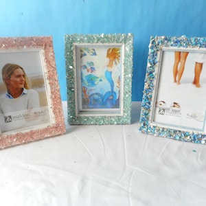 May include: Three decorative photo frames, each with a different color scheme. The frames are encrusted with small, sparkling pieces. The frames hold 4x6 inch photos. One frame is pink, one is green, and one is blue.