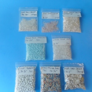 May include: Eight clear plastic resealable bags containing various craft materials including white shell crush, pink shell crush, pale blue stone chips, white stone chips, white pebbles, faux flower buds, and medium shell crush with handwritten labels.