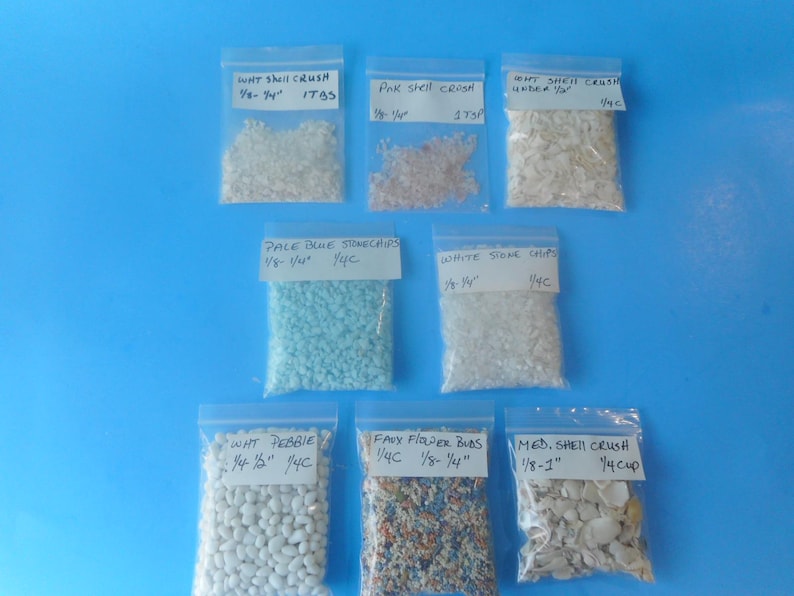 May include: Eight clear plastic resealable bags containing various craft materials including white shell crush, pink shell crush, pale blue stone chips, white stone chips, white pebbles, faux flower buds, and medium shell crush with handwritten labels.