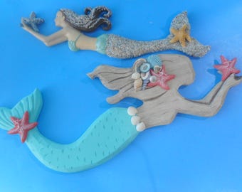 May include: Two mermaid figurines with starfish accents. One mermaid has a light blue tail and a red starfish. The other mermaid has a sand-textured tail and a yellow starfish. Both mermaids are on a blue background.