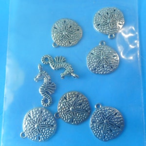 May include: A collection of silver-toned metal charms featuring six circular sand dollar shapes with textured surfaces and two seahorse shapes, each equipped with a small loop at the top for attachment, contained within a clear plastic bag.