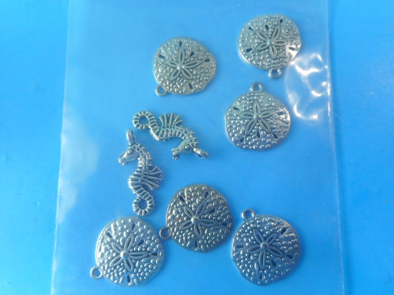 May include: A collection of silver-toned metal charms featuring six circular sand dollar shapes with textured surfaces and two seahorse shapes, each equipped with a small loop at the top for attachment, contained within a clear plastic bag.