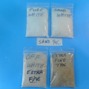 May include: Four clear plastic resealable bags filled with sand. The bags are labeled with handwritten text: PURE WHITE, SAND WHITE, OFF WHITE EXTRA FINE, and EXTRA FINE TAN. A small white paper label between the bags reads SAND 1/4C.