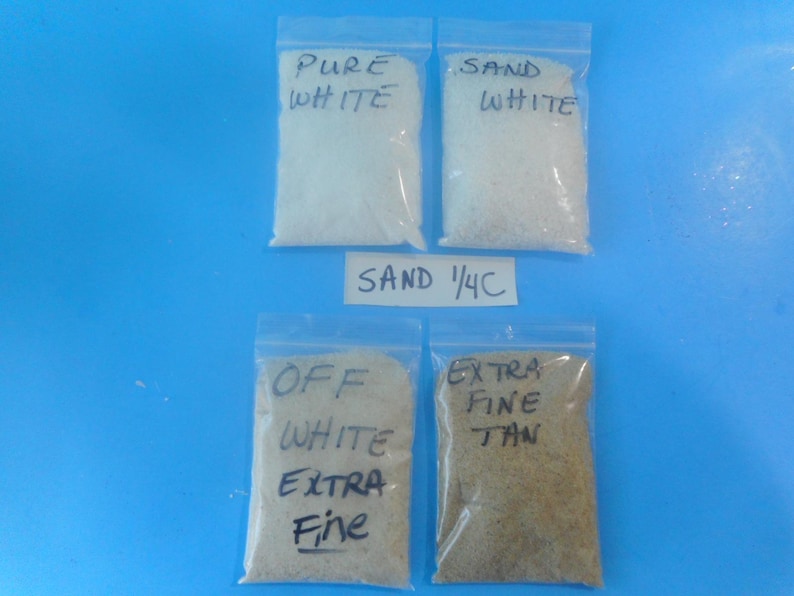 May include: Four clear plastic resealable bags filled with sand. The bags are labeled with handwritten text: PURE WHITE, SAND WHITE, OFF WHITE EXTRA FINE, and EXTRA FINE TAN. A small white paper label between the bags reads SAND 1/4C.