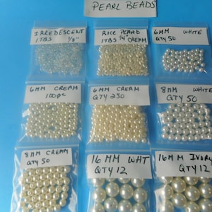 May include: Nine clear plastic bags containing various pearl beads, labeled with sizes and quantities. Labels include Iridescent 1/8 inch, Rice Pearl 1/4 inch, 6mm White, 6mm Cream 100pc, 6mm Cream 250, 8mm White, 8mm Cream 50, 16mm White, and 16mm Ivory.