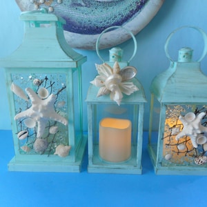 May include: Three teal blue lanterns decorated with seashells and netting. The lanterns are lit with candles and are perfect for a beach-themed home decor.