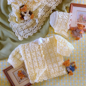 May include: A set of crocheted baby clothes in a cream color, including a cardigan, a hat, and a blanket. The set is laid out on a green and yellow patterned fabric.