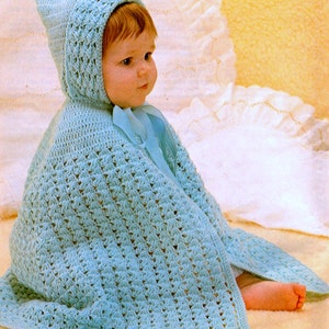 May include: A baby wearing a light blue crocheted hooded cape with a matching ribbon tie. The cape has a textured pattern and covers the baby's shoulders and arms. The baby is sitting on a soft, light-colored surface.