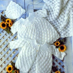 May include: A white knitted baby outfit set including a hat, a dress, and a cardigan. The set is made with a delicate lace pattern and features a button closure on the cardigan.