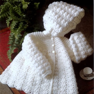 May include: White crocheted baby jacket with a hood and button fastening. The jacket has a textured, puffy design.