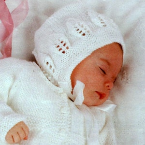 May include: A white knitted baby bonnet with a delicate lace pattern. The bonnet is worn by a sleeping baby.