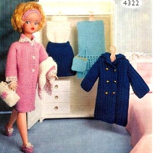 Puede incluir: Una muñeca con un abrigo de punto rosa y una bufanda de punto blanca. La muñeca está de pie frente a una cómoda blanca con tres cajones. Hay tres prendas de punto colgadas en ganchos sobre la cómoda: una falda azul, un vestido azul claro y un abrigo azul oscuro.