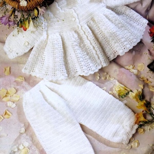 May include: A white crocheted baby outfit with a ruffled cardigan and matching pants. The outfit is laid out on a pink and white floral background.