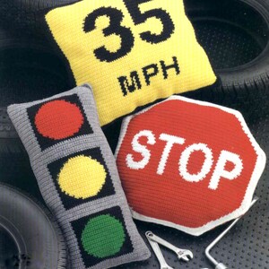 K&ouml;nnte beinhalten: Drei geh&auml;kelte Kissen in Form von Verkehrsschildern. Das erste Kissen ist gelb mit schwarzem Text, der "35 MPH" lautet. Das zweite Kissen ist grau mit roten, gelben und gr&uuml;nen Kreisen, die einem Verkehrsampel &auml;hneln. Das dritte Kissen ist rot mit wei&szlig;em Text, der "STOP" lautet.