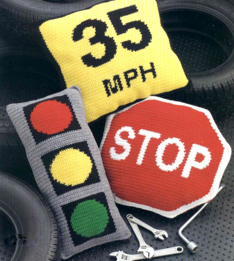 K&ouml;nnte beinhalten: Drei geh&auml;kelte Kissen in Form von Verkehrsschildern. Das erste Kissen ist gelb mit schwarzem Text, der "35 MPH" lautet. Das zweite Kissen ist grau mit roten, gelben und gr&uuml;nen Kreisen, die einem Verkehrsampel &auml;hneln. Das dritte Kissen ist rot mit wei&szlig;em Text, der "STOP" lautet.