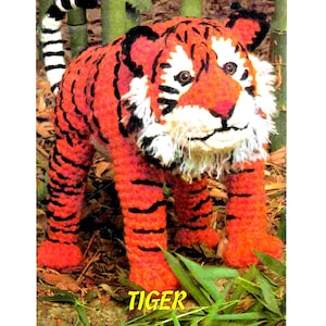 Puede incluir: Un juguete de peluche de tigre tejido a crochet con rayas naranjas y negras. El tigre está de pie sobre una cama de hojas verdes y ramitas marrones. La palabra "TIGER" está impresa en letras negras debajo del tigre.