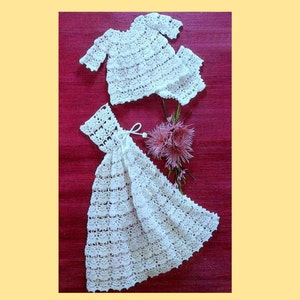 May include: A white crocheted baby dress and matching top set. The dress has a long, flowing skirt and a fitted bodice. The top has short sleeves and a round neckline. The set is laid out on a red background.