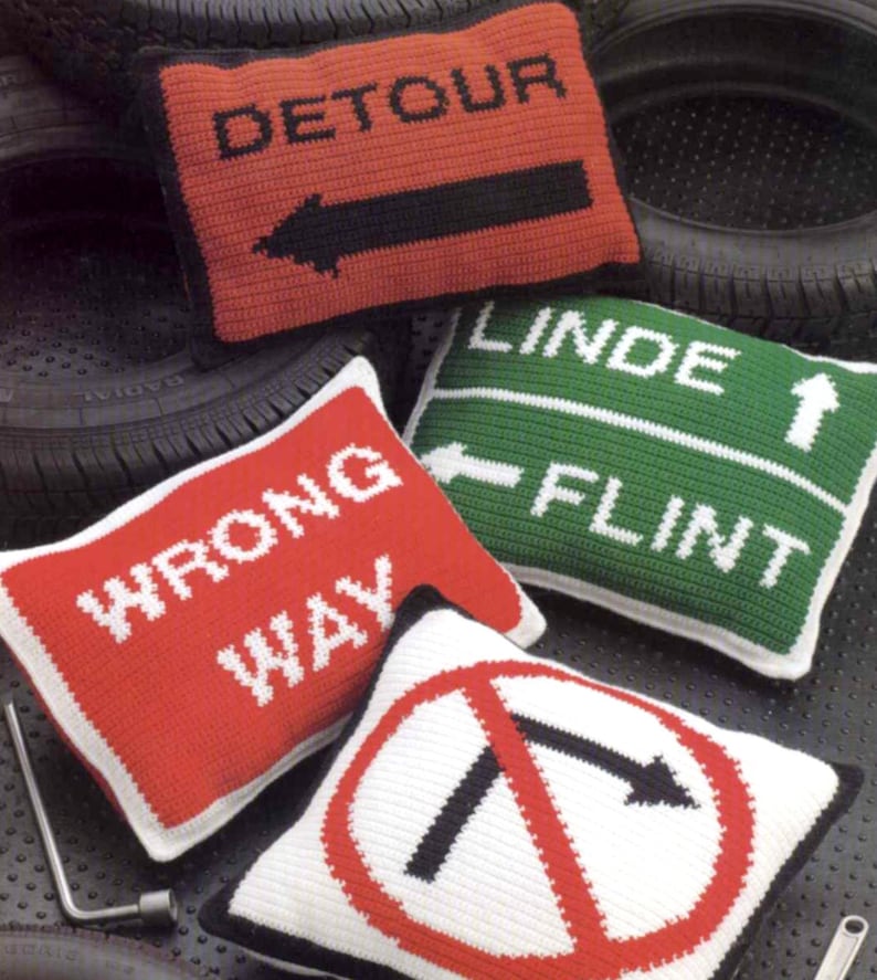 K&ouml;nnte beinhalten: Vier geh&auml;kelte Kissen in Form von Verkehrsschildern. Die Schilder zeigen "Umleitung", "Falsche Richtung", "Linde Flint" und ein Schild mit einem Rechtsabbiegeverbot.