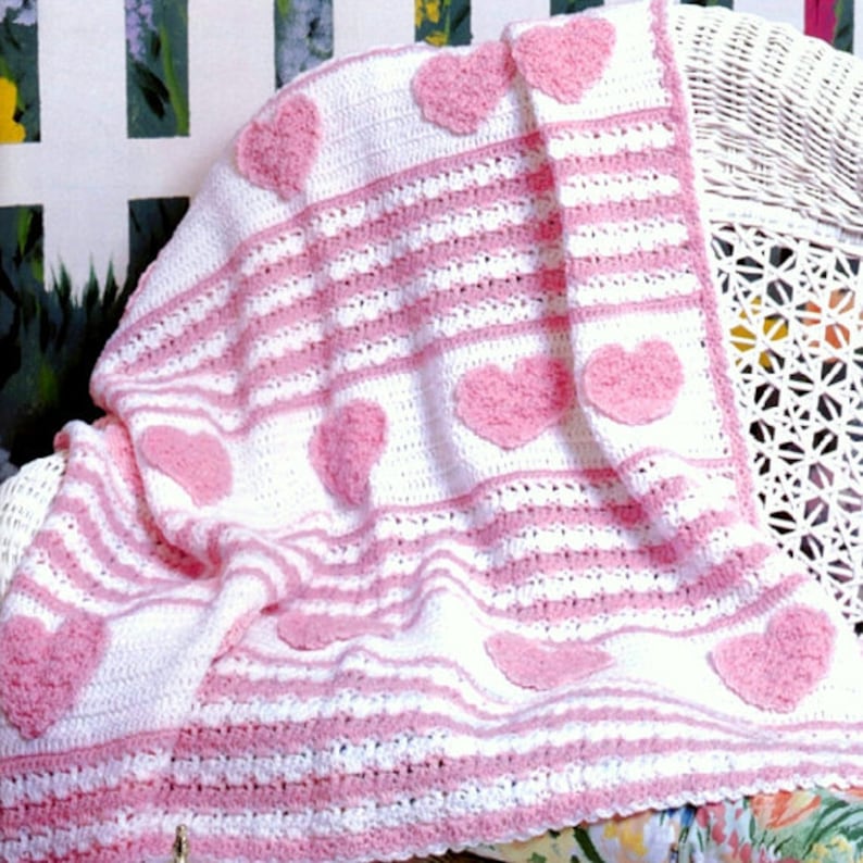 May include: A handmade crochet blanket in white and pink. The blanket features alternating stripes of white and pink, with pink heart appliqu&eacute;s scattered across the surface. The blanket is draped over a white wicker chair.
