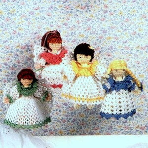 May include: Four crocheted dolls wearing different colored dresses. The dolls are all wearing white dresses with different colored accents. The dolls are standing on a floral patterned background.
