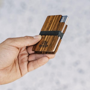 Puede incluir: Un porta tarjetas de madera con una banda el&aacute;stica negra. El porta tarjetas tiene un patr&oacute;n de veta de madera marr&oacute;n y el texto "Woodchuck" est&aacute; grabado en la parte delantera. El porta tarjetas tiene una tarjeta VISA insertada.