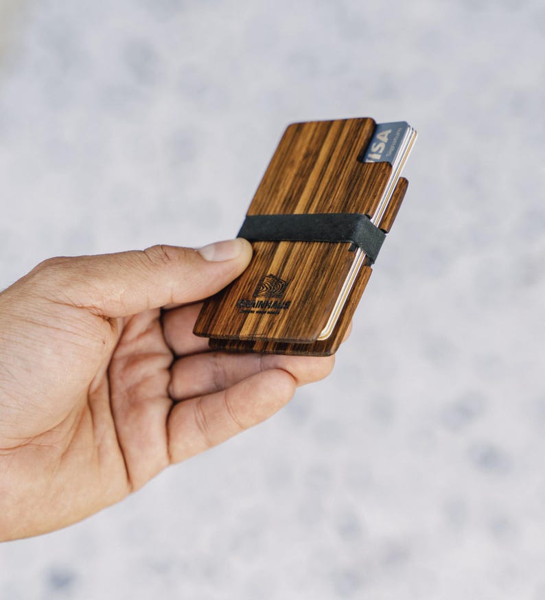 Puede incluir: Un porta tarjetas de madera con una banda el&aacute;stica negra. El porta tarjetas tiene un patr&oacute;n de veta de madera marr&oacute;n y el texto "Woodchuck" est&aacute; grabado en la parte delantera. El porta tarjetas tiene una tarjeta VISA insertada.
