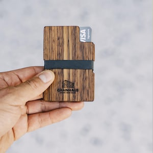 Puede incluir: Una cartera de madera con una banda el&aacute;stica negra y una tarjeta de cr&eacute;dito VISA Signature. La cartera tiene un patr&oacute;n de veta de madera y el texto "GRAINHAUS" est&aacute; grabado en la parte delantera.