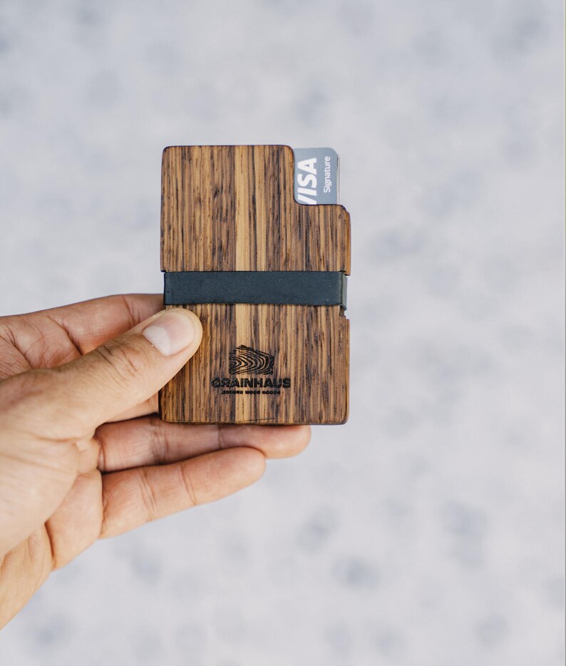 Puede incluir: Una cartera de madera con una banda el&aacute;stica negra y una tarjeta de cr&eacute;dito VISA Signature. La cartera tiene un patr&oacute;n de veta de madera y el texto "GRAINHAUS" est&aacute; grabado en la parte delantera.