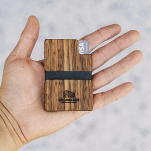 Puede incluir: Una cartera de madera con una banda el&aacute;stica negra y una tarjeta VISA Signature. La cartera tiene un patr&oacute;n de veta de madera y el texto "GRAINHAUS" est&aacute; grabado en la parte delantera.