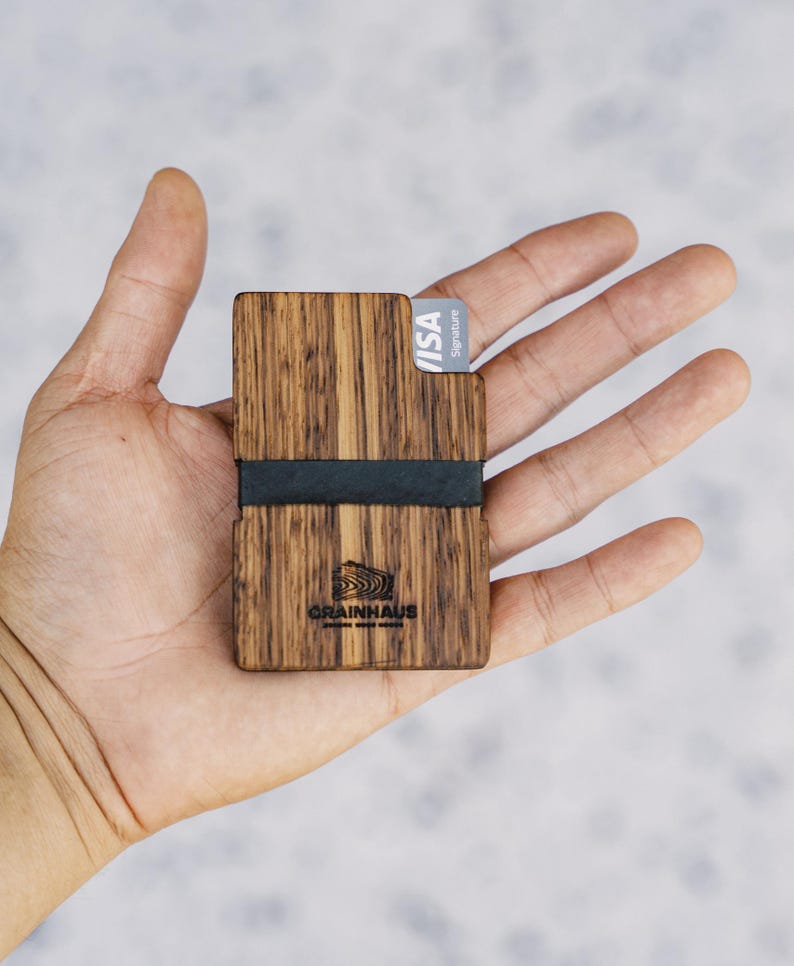 Puede incluir: Una cartera de madera con una banda el&aacute;stica negra y una tarjeta VISA Signature. La cartera tiene un patr&oacute;n de veta de madera y el texto "GRAINHAUS" est&aacute; grabado en la parte delantera.