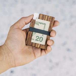 Puede incluir: Una cartera de madera con una banda el&aacute;stica negra que sostiene un billete de veinte euros y una tarjeta de cr&eacute;dito.