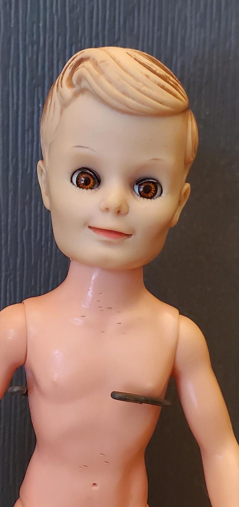 May include: A plastic doll with light brown hair and brown eyes. The doll is wearing a nude-coloured body and has a small belly button.