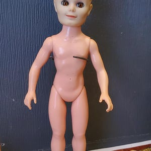 May include: A nude plastic doll with blonde hair and brown eyes. The doll is standing on a grey circular base.