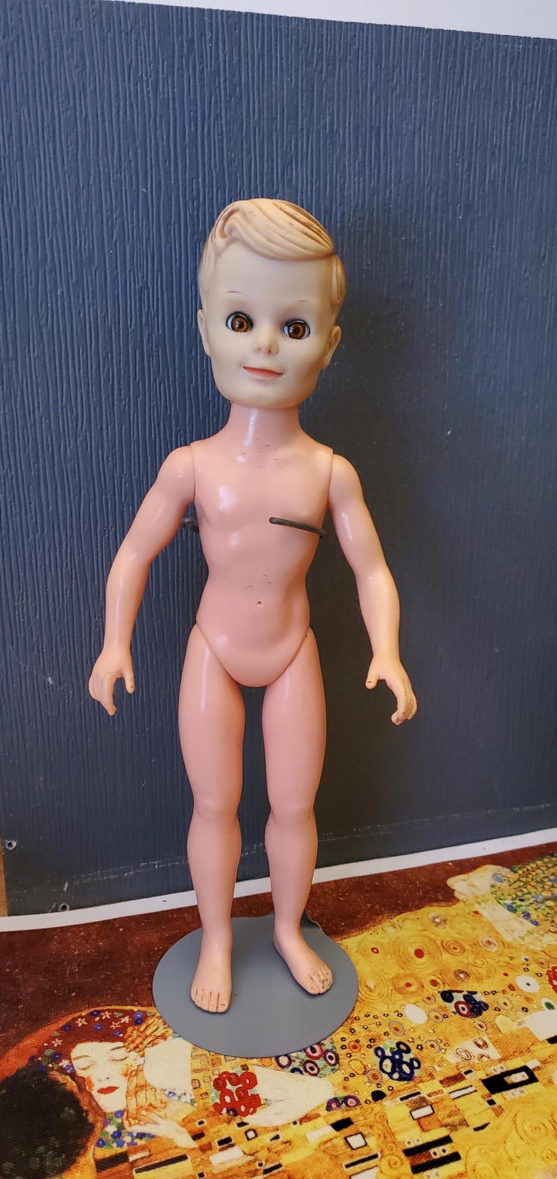 May include: A nude plastic doll with blonde hair and brown eyes. The doll is standing on a grey circular base.