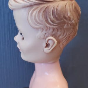 May include: A vintage plastic doll with a sculpted hairstyle and a light pink body. The doll is wearing a brown and white striped outfit.
