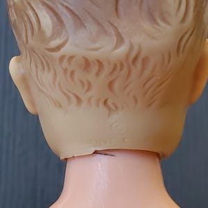 May include: The back of a plastic doll's head with a moulded hairstyle. The doll has a neck seam and a "WHED" trademark on the back of the neck.