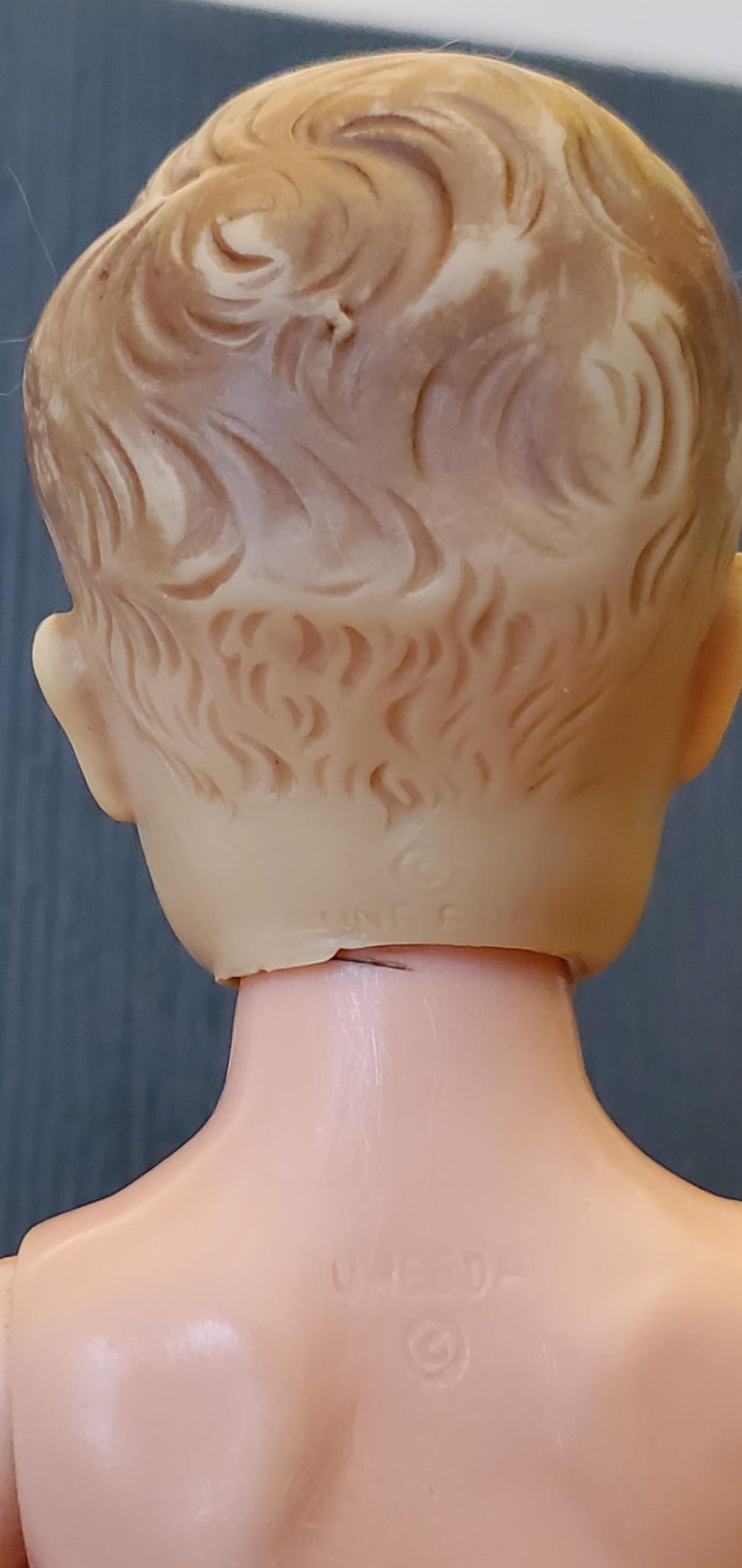 May include: The back of a plastic doll's head with a moulded hairstyle. The doll has a neck seam and a "WHED" trademark on the back of the neck.