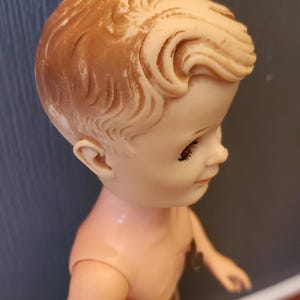 May include: A vintage plastic doll with a sculpted hairstyle and brown eyes. The doll is wearing a beige cloth towel around its waist.