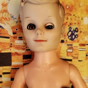 May include: A vintage plastic doll with blonde hair and brown eyes. The doll is wearing a brown and white striped shirt and brown trousers. The doll is missing one eye.