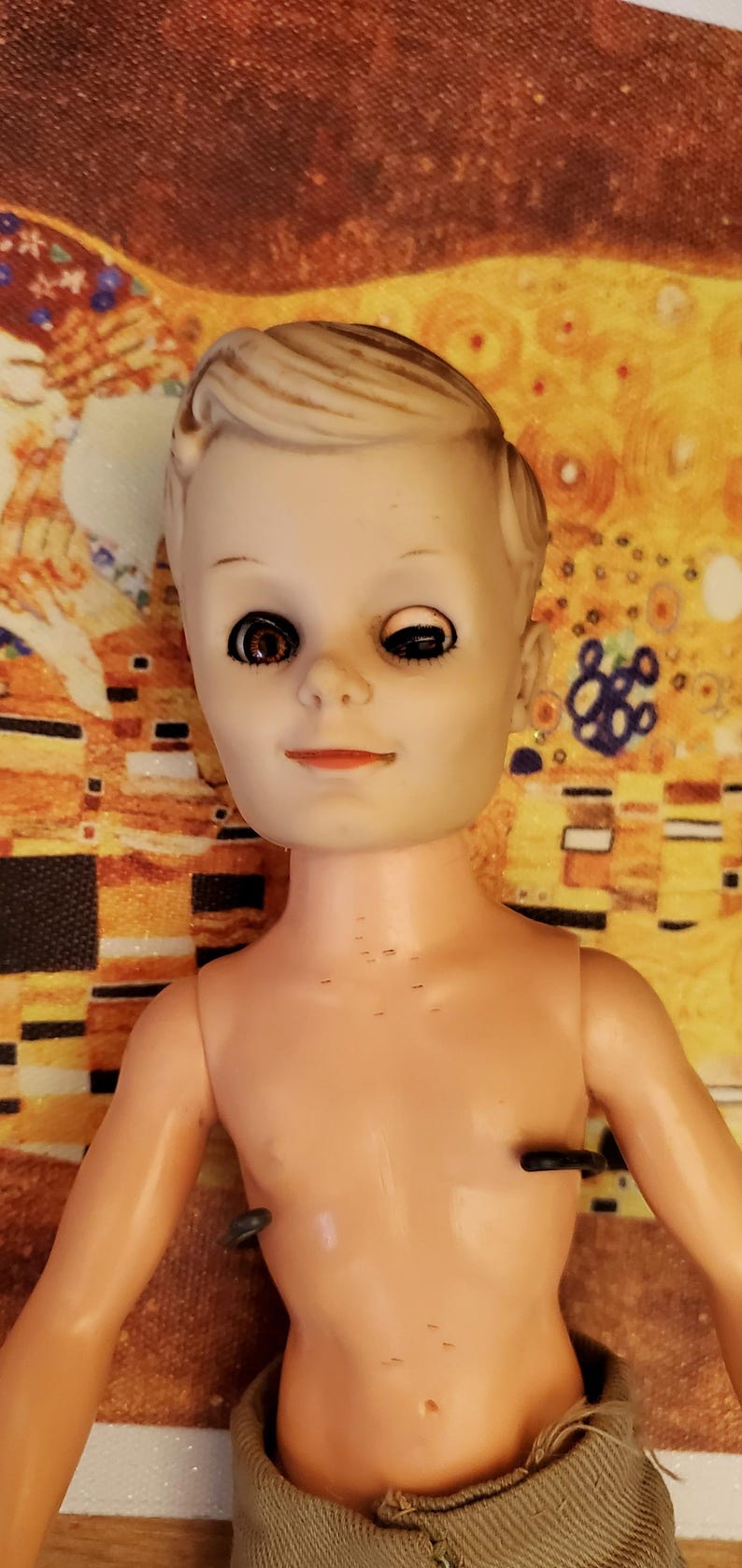 May include: A vintage plastic doll with blonde hair and brown eyes. The doll is wearing a brown and white striped shirt and brown trousers. The doll is missing one eye.
