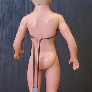 May include: A nude plastic doll with blonde hair standing on a grey metal doll stand. The doll is posed with its arms at its sides and its legs slightly apart.