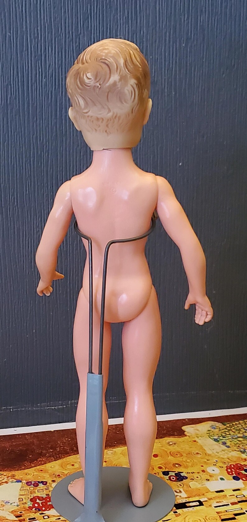 May include: A nude plastic doll with blonde hair standing on a grey metal doll stand. The doll is posed with its arms at its sides and its legs slightly apart.