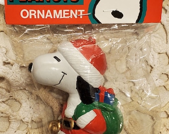 May include: A white Peanuts Snoopy Christmas ornament. Snoopy is dressed as Santa Claus, wearing a red hat and a red and white suit. He is carrying a green bag with a blue ribbon. The ornament is packaged in a clear plastic bag with a red and white Peanuts ornament label.