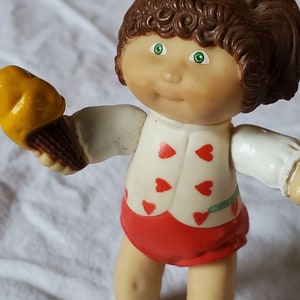May include: A plastic doll with brown hair in pigtails, wearing a white shirt with red hearts, red shorts, and white shoes. The doll is holding a yellow and brown ice cream cone.